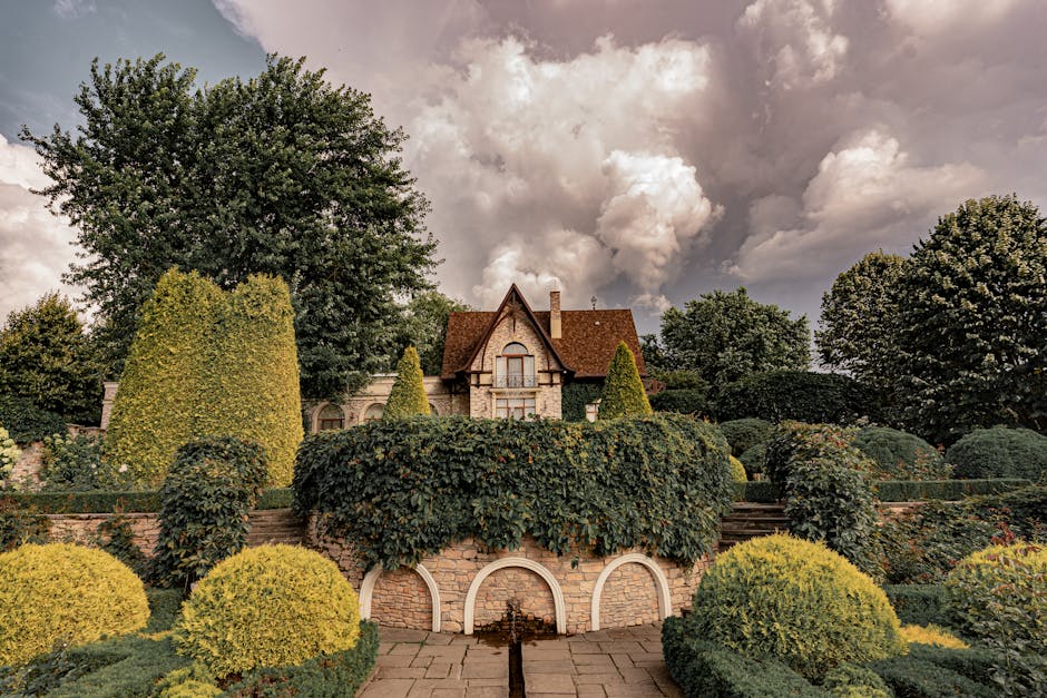 Elegant manor house framed by topiary and lush garden with dramatic cloudy sky backdrop.